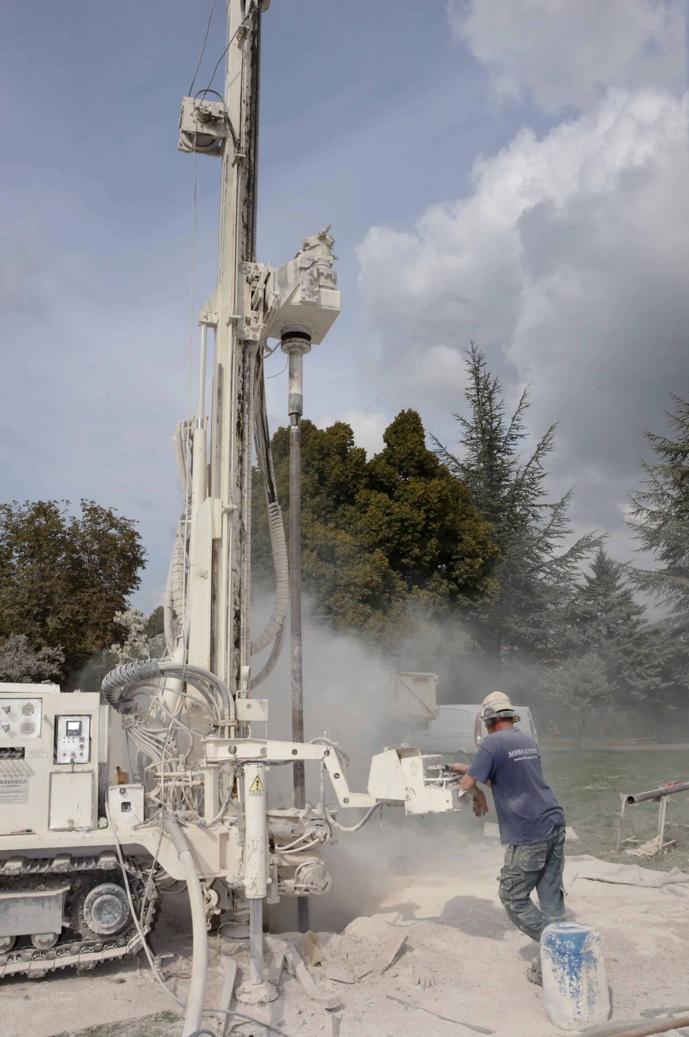 A borehole drilling rig at work with dust rising around the drill pipe while a driller in a hard hat guides the rod