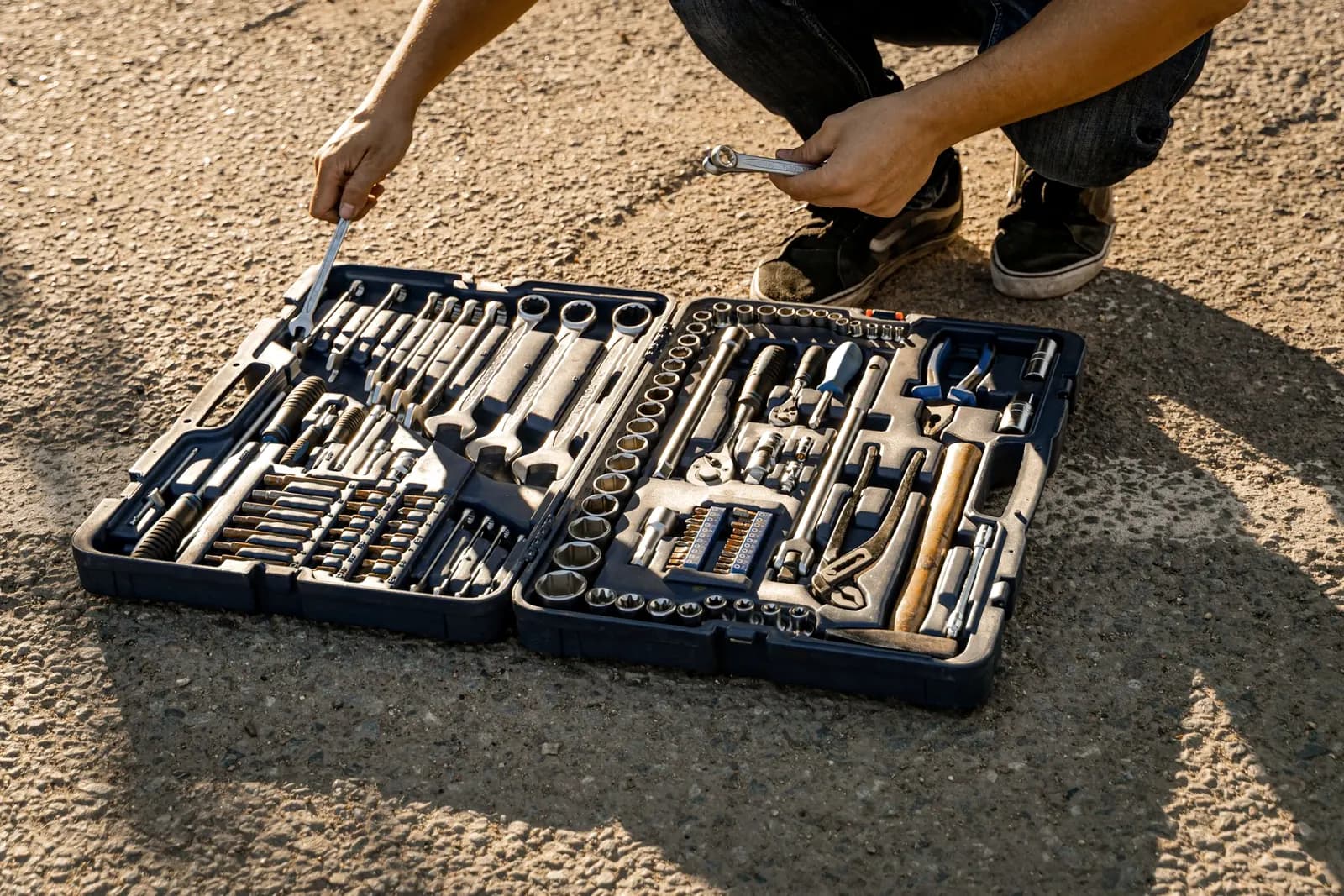 A technician crouching over an open toolbox of spanners and sockets on sunlit ground