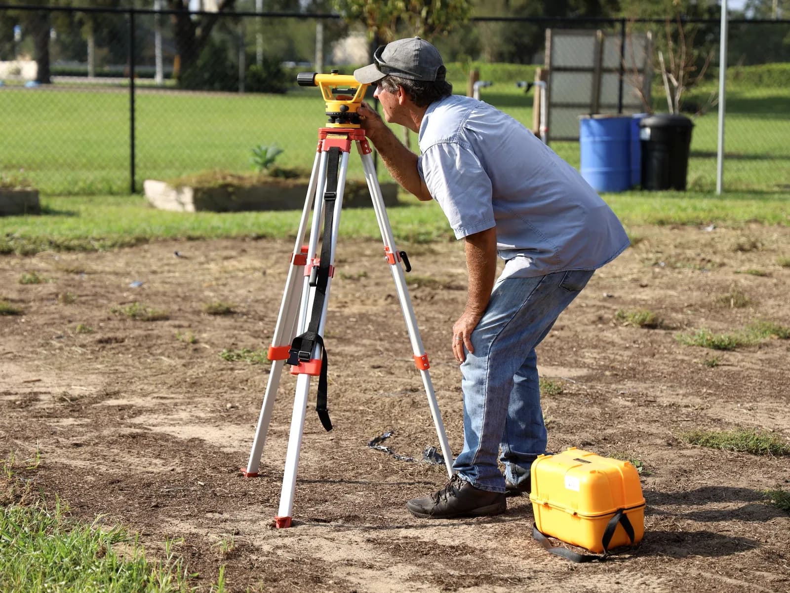 A surveyor in a cap sighting through a level on a tripod in an open yard, with a yellow equipment case at his feet